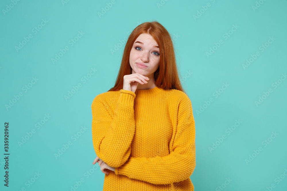 Confused puzzled young redhead woman girl in yellow sweater posing isolated on blue turquoise wall background studio portrait. People lifestyle concept. Mock up copy space. Put hand prop up on chin. Confused puzzled young redhead woman girl in yellow sweater posing isolated on blue turquoise wall background studio portrait. People lifestyle concept. Mock up copy space. Put hand prop up on chin.