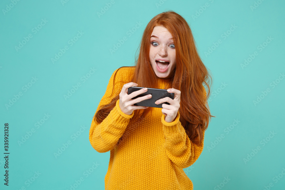 Excited young redhead woman girl in yellow sweater posing isolated on blue turquoise background studio portrait. People emotions lifestyle concept. Mock up copy space. Play game with mobile phone. Excited young redhead woman girl in yellow sweater posing isolated on blue turquoise background studio portrait. People emotions lifestyle concept. Mock up copy space. Play game with mobile phone.