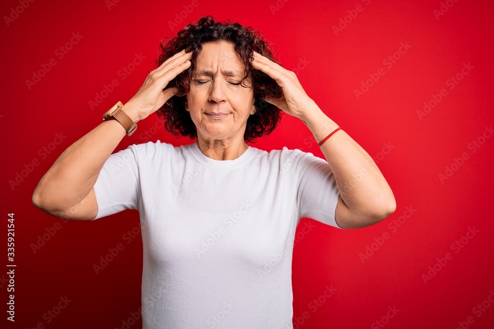 Middle age beautiful curly hair woman wearing casual t-shirt over isolated red background suffering from headache desperate and stressed because pain and migraine. Hands on head. Middle age beautiful curly hair woman wearing casual t-shirt over isolated red background suffering from headache desperate and stressed because pain and migraine. Hands on head.