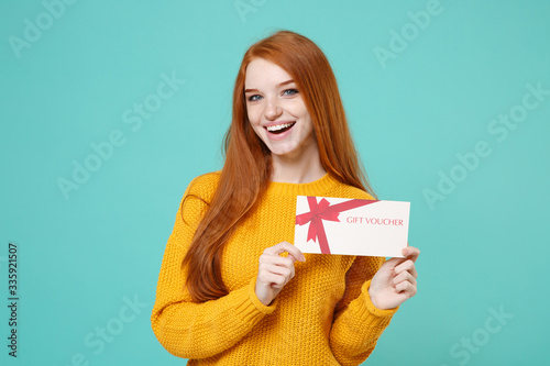 Cheerful young redhead woman girl in yellow knitted sweater posing isolated on blue turquoise background studio portrait. People emotions lifestyle concept. Mock up copy space. Hold gift certificate.