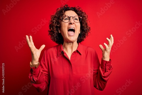 Middle age beautiful curly hair woman wearing casual shirt and glasses over red background crazy and mad shouting and yelling with aggressive expression and arms raised. Frustration concept.