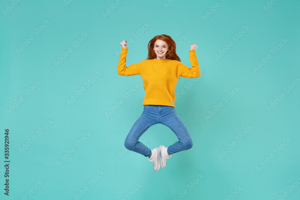 Funny young redhead woman girl in yellow knitted sweater posing isolated on blue turquoise background studio portrait. People emotions lifestyle concept. Mock up copy space. Jumping clenching fists. Funny young redhead woman girl in yellow knitted sweater posing isolated on blue turquoise background studio portrait. People emotions lifestyle concept. Mock up copy space. Jumping clenching fists.