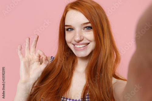 Close up of smiling young redhead girl in plaid dress posing isolated on pastel pink background. People lifestyle concept. Mock up copy space. Doing selfie shot on mobile phone, showing OK gesture.