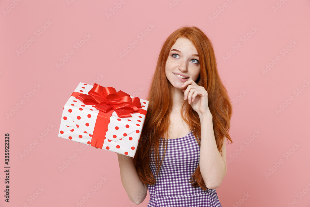 Pensive redhead girl in plaid dress isolated on pastel pink background. Valentine's Day Women's Day birthday holiday concept. Hold white red present box with gift ribbon bow, put hand prop up on chin. Pensive redhead girl in plaid dress isolated on pastel pink background. Valentine's Day Women's Day birthday holiday concept. Hold white red present box with gift ribbon bow, put hand prop up on chin.