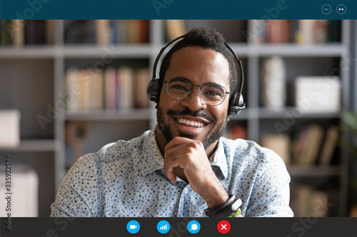 Head shot African man wear headset sit at desk look at cam use videocall conferencing app, pc screen view. Company consultant, teacher portrait, easy e-learn, distant communication with client concept