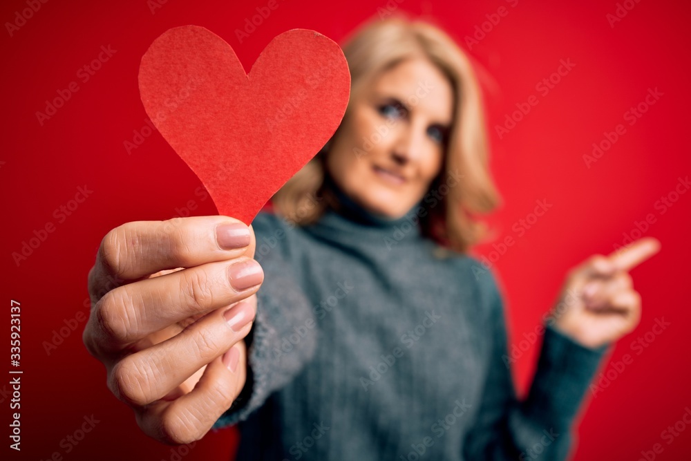 Middle age beautiful blonde romantic woman holding red paper heart celebrating valentine day very happy pointing with hand and finger to the side Middle age beautiful blonde romantic woman holding red paper heart celebrating valentine day very happy pointing with hand and finger to the side