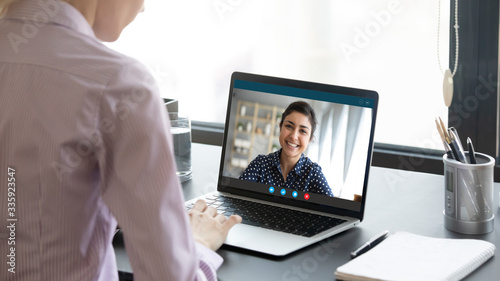 Indian girl communicate with friend on-line by video call, pc screen view over female shoulder. Mental health expert online therapy, colleagues work on common project use videoconferencing app concept photo