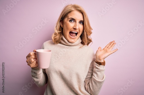 Middle age beautiful blonde woman drinking pink mug of coffee over isolated background very happy and excited, winner expression celebrating victory screaming with big smile and raised hands
