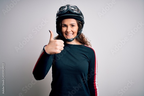 Beautiful motorcyclist woman with curly hair wearing moto helmet over white background doing happy thumbs up gesture with hand. Approving expression looking at the camera showing success.