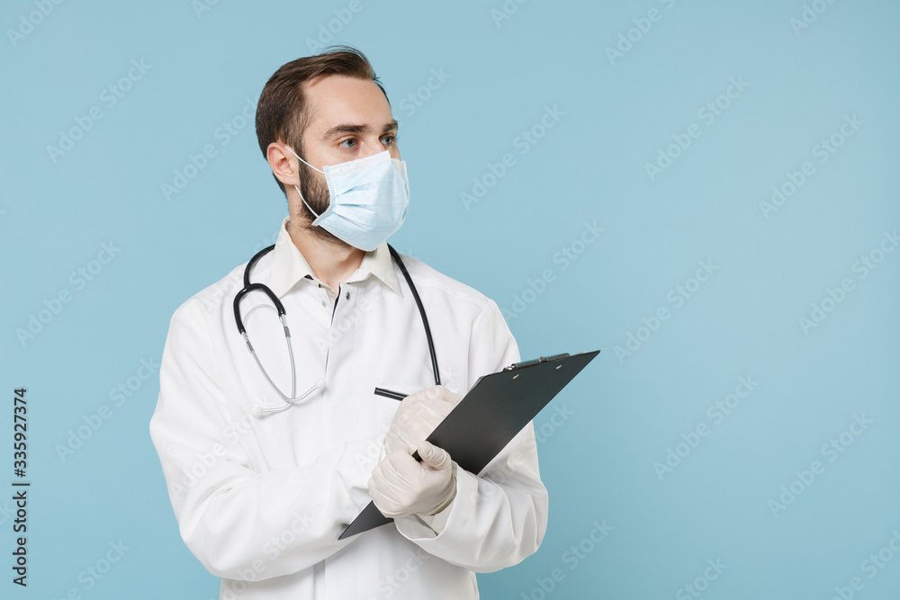 Male doctor man in medical gown face mask gloves isolated on blue background. Epidemic pandemic coronavirus 2019-ncov sars covid-19 flu virus concept. Hold clipboard with medical document health card.
