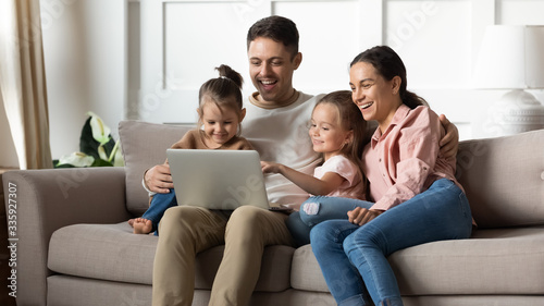 Happy married couple resting on sofa with adorable little children daughters, watching funny movies on computer at home. Laughing family playing online game or shopping together in living room.