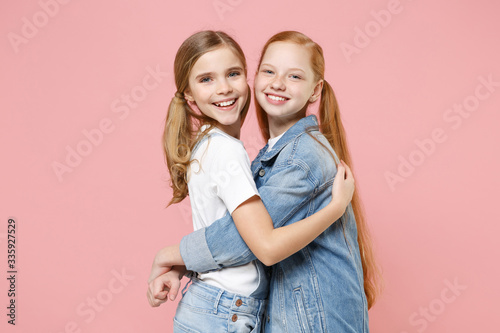 Smiling funny little kids girls 12-13 years old in white t-shirt, denim clothes isolated on pastel pink background children studio portrait. Childhood lifestyle concept. Mock up copy space. Hugging.