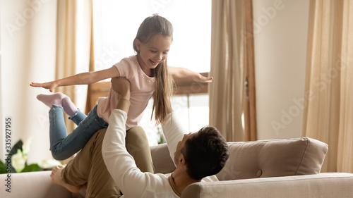Happy little preschool girl making airplane, practicing yoga with cheerful father on couch. Young dad holding on straight hands small child daughter, involved in funny family activity on sofa at home.