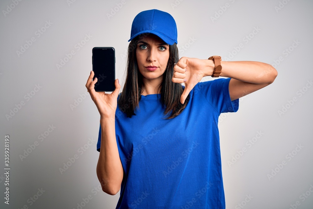 Young delivery woman with blue eyes wearing cap holding smartphone with angry face, negative sign showing dislike with thumbs down, rejection concept