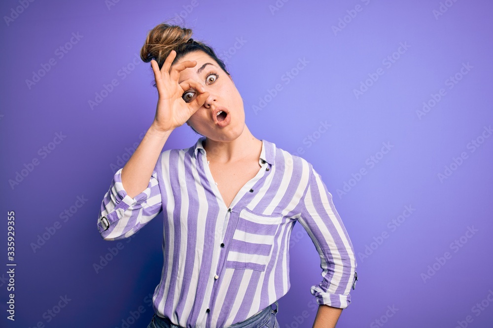 Young beautiful blonde woman wearing casual striped shirt standing over purple background doing ok gesture shocked with surprised face, eye looking through fingers. Unbelieving expression. Young beautiful blonde woman wearing casual striped shirt standing over purple background doing ok gesture shocked with surprised face, eye looking through fingers. Unbelieving expression.