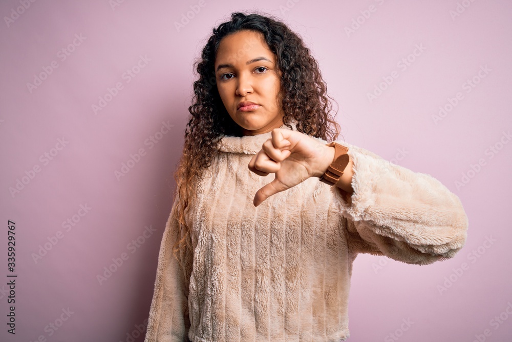 Young beautiful woman with curly hair wearing casual sweater standing over pink background looking unhappy and angry showing rejection and negative with thumbs down gesture. Bad expression. Young beautiful woman with curly hair wearing casual sweater standing over pink background looking unhappy and angry showing rejection and negative with thumbs down gesture. Bad expression.