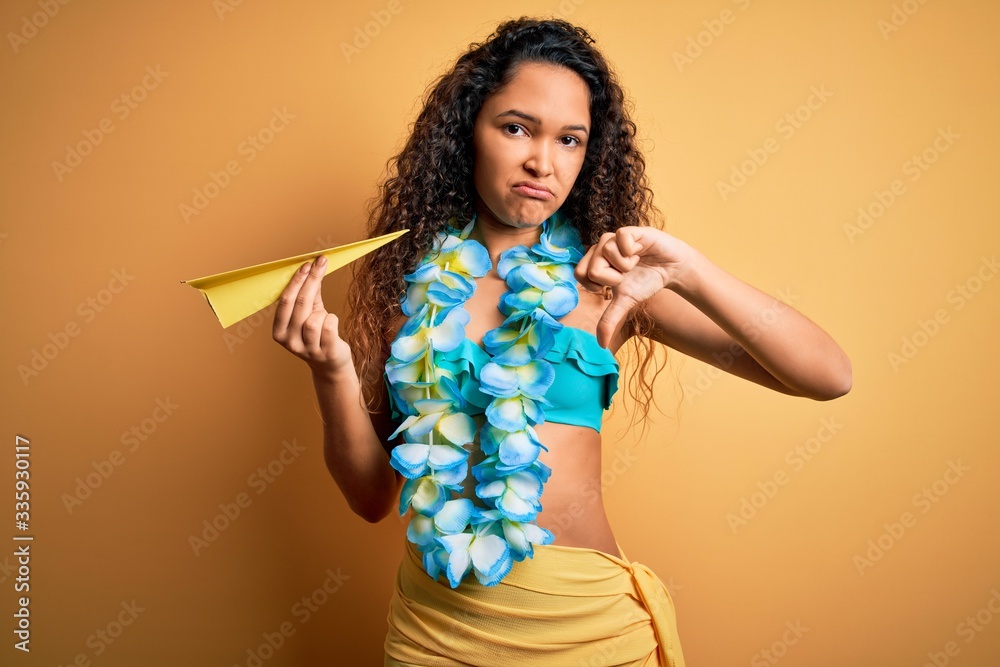 Young beautiful american woman on vacation wearing bikini holding paper airplane with angry face, negative sign showing dislike with thumbs down, rejection concept Young beautiful american woman on vacation wearing bikini holding paper airplane with angry face, negative sign showing dislike with thumbs down, rejection concept