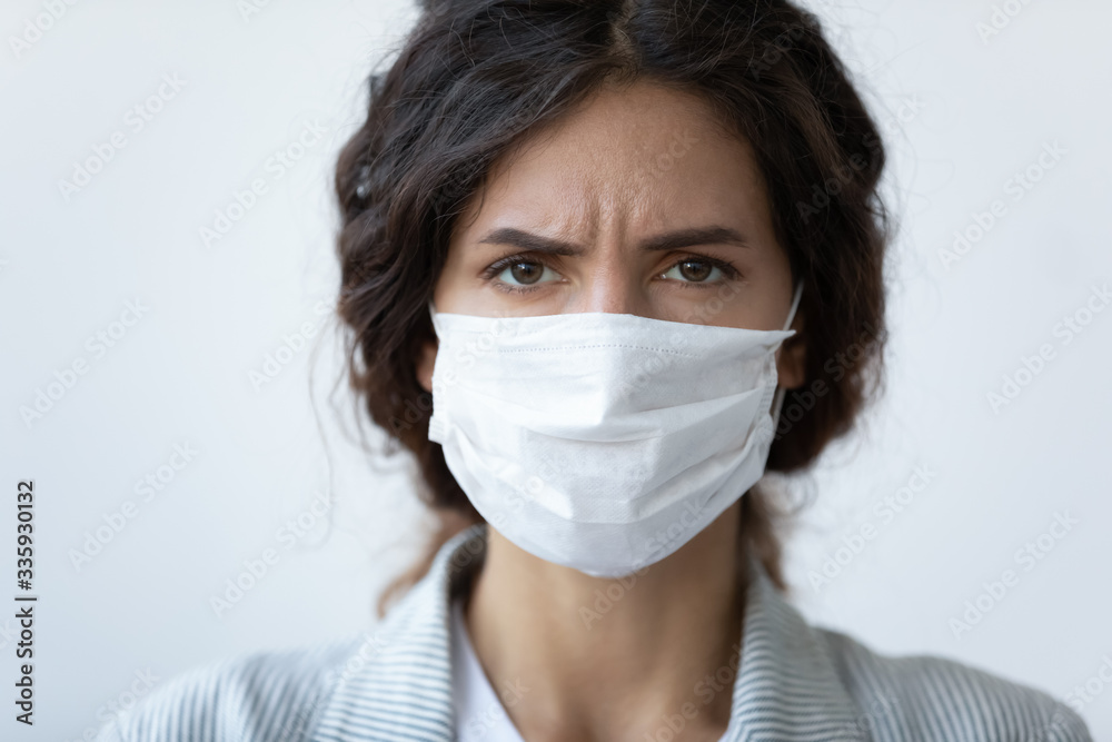 Head shot close up portrait frowning stressed unhappy young woman wearing medical facial mask, isolated on blue studio background. Worrying lady with covered mouth and nose looking at camera. Head shot close up portrait frowning stressed unhappy young woman wearing medical facial mask, isolated on blue studio background. Worrying lady with covered mouth and nose looking at camera.