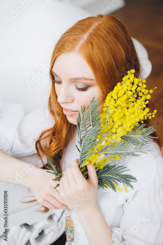 Portrait of a red-haired beautiful girl with a Mimosa in a long white dress