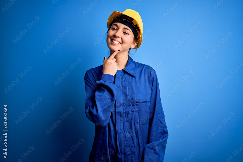 Fototapeta premium Young beautiful worker woman with blue eyes wearing security helmet and uniform looking confident at the camera with smile with crossed arms and hand raised on chin. Thinking positive.