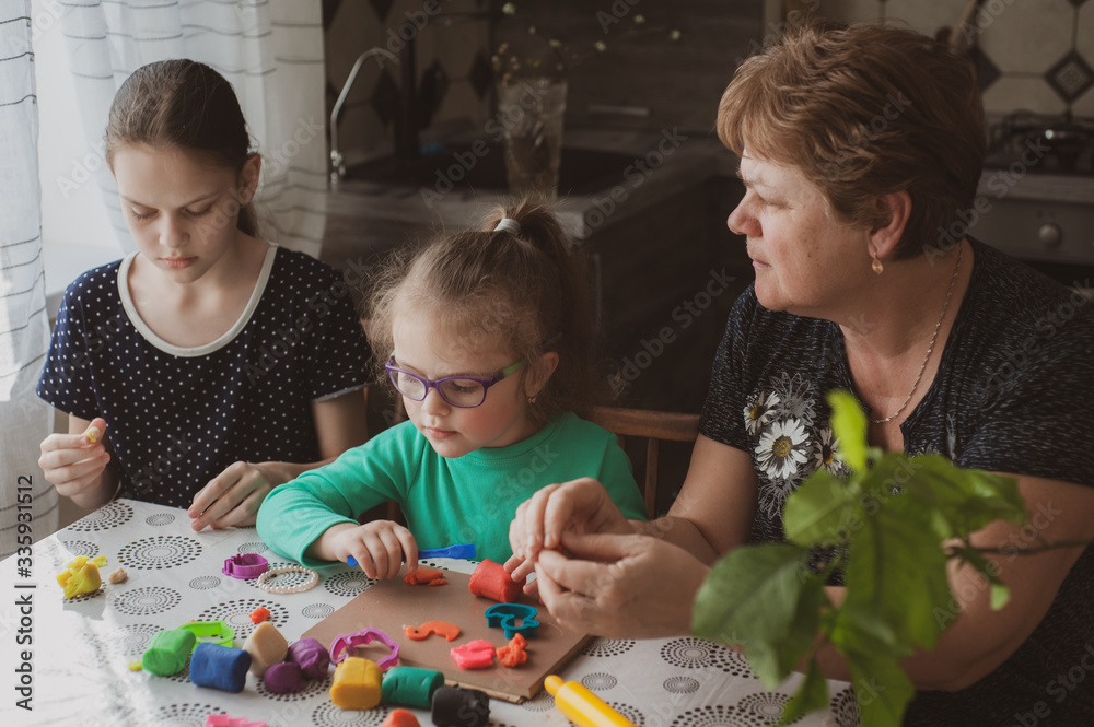Fototapeta premium An adult woman and two little girls make plasticine figurines at home. Grandmother and granddaughters are sitting at home. Family Values Concept