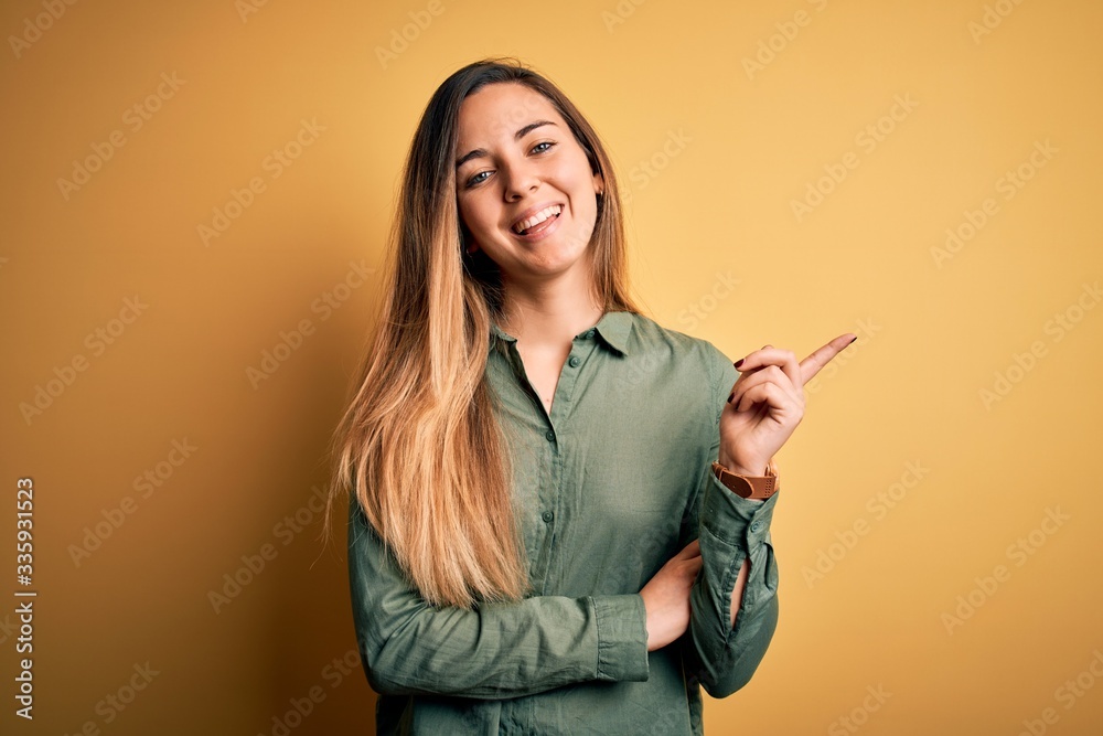 Young beautiful blonde woman with blue eyes wearing green shirt over yellow background with a big smile on face, pointing with hand and finger to the side looking at the camera. Young beautiful blonde woman with blue eyes wearing green shirt over yellow background with a big smile on face, pointing with hand and finger to the side looking at the camera.