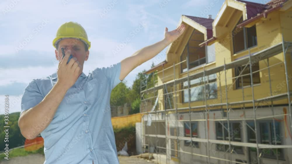 SLOW MOTION, CLOSE UP: Angry Caucasian contractor shouts at his co-worker over the walkie talkie while standing outside of a house under construction. Architect gets into an argument with contractors.