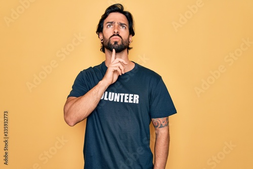 Young handsome hispanic volunteer man wearing volunteering t-shirt as social care Thinking concentrated about doubt with finger on chin and looking up wondering