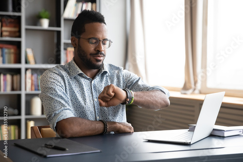 African American young male employee in glasses check time on hand watch waiting for work day to be over, biracial man worker sit at desk use smartwatch at workplace, time management concept
