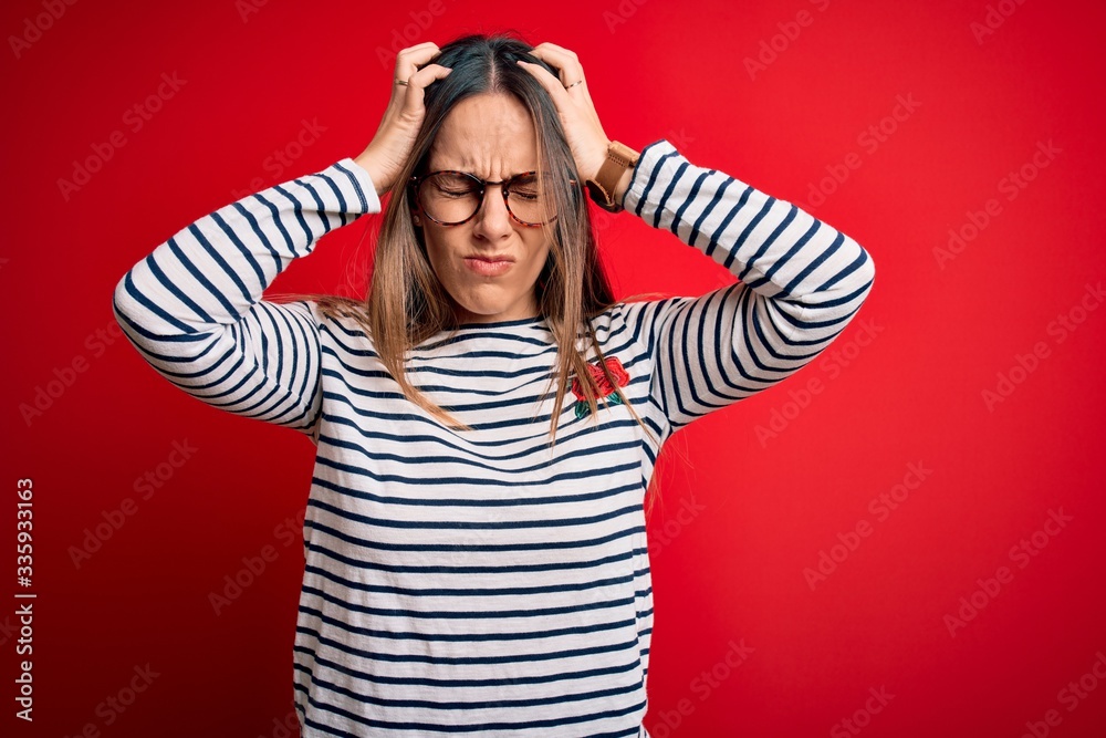 Young beautiful blonde woman with blue eyes wearing glasses standing over red background suffering from headache desperate and stressed because pain and migraine. Hands on head. Young beautiful blonde woman with blue eyes wearing glasses standing over red background suffering from headache desperate and stressed because pain and migraine. Hands on head.