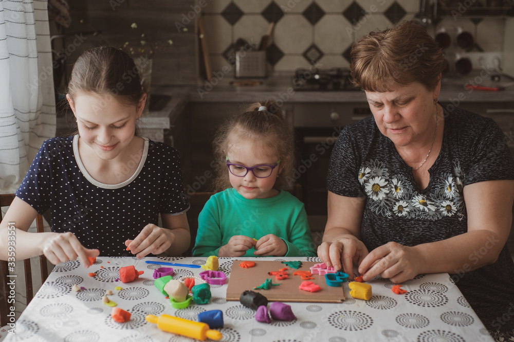 Fototapeta premium An adult woman and two little girls make plasticine figurines at home. Grandmother and granddaughters are sitting at home. Family Values Concept