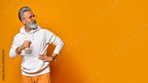 Bearded grandpa in white hoodie, pants and sunglasses, bracelets. Put his hand on hip, posing on orange background. Close up