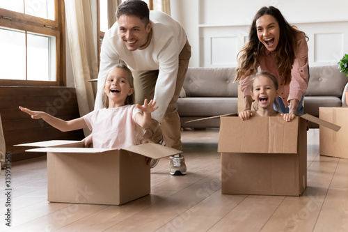 Full length overjoyed little sisters sitting in big cardboard boxes, playing with happy parents in new home. Excited laughing family couple having fun together with adorable daughters in living room.