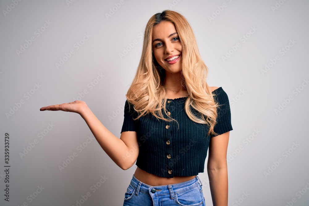 Young beautiful blonde woman wearing casual t-shirt standing over isolated white background smiling cheerful presenting and pointing with palm of hand looking at the camera. Young beautiful blonde woman wearing casual t-shirt standing over isolated white background smiling cheerful presenting and pointing with palm of hand looking at the camera.
