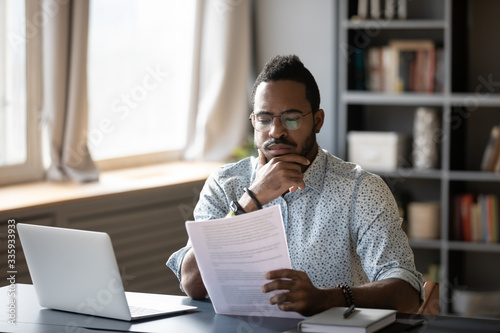 Pensive African American man sit at desk working on laptop reading paper document thinking or analyzing, thoughtful biracial male worker consider paperwork agreement at office or home workplace