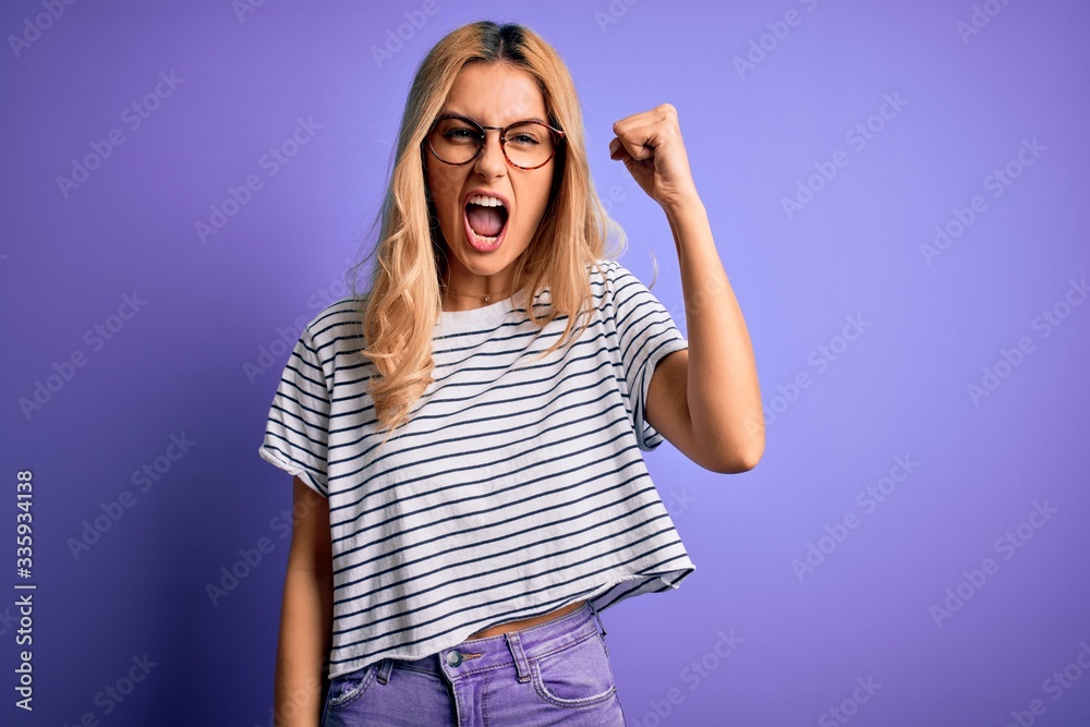 Fototapeta premium Young beautiful blonde woman wearing striped t-shirt and glasses over purple background angry and mad raising fist frustrated and furious while shouting with anger. Rage and aggressive concept.