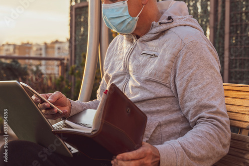 Man with a mask sitting on a wooden bench outdoors working with a laptop a telephone and an agenda in a time of confinement due to the covid 19 pandemic