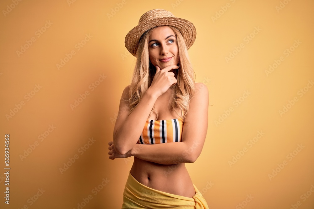 Young beautiful blonde woman on vacation wearing bikini and hat over yellow background with hand on chin thinking about question, pensive expression. Smiling with thoughtful face. Doubt concept. Young beautiful blonde woman on vacation wearing bikini and hat over yellow background with hand on chin thinking about question, pensive expression. Smiling with thoughtful face. Doubt concept.