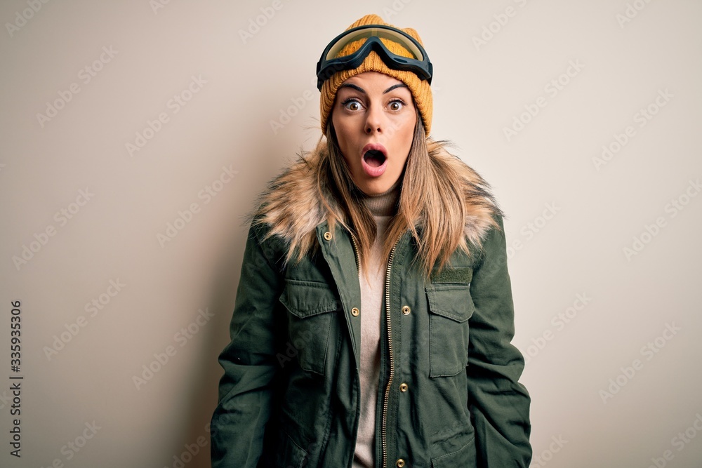 Young brunette skier woman wearing snow clothes and ski goggles over white background afraid and shocked with surprise and amazed expression, fear and excited face.