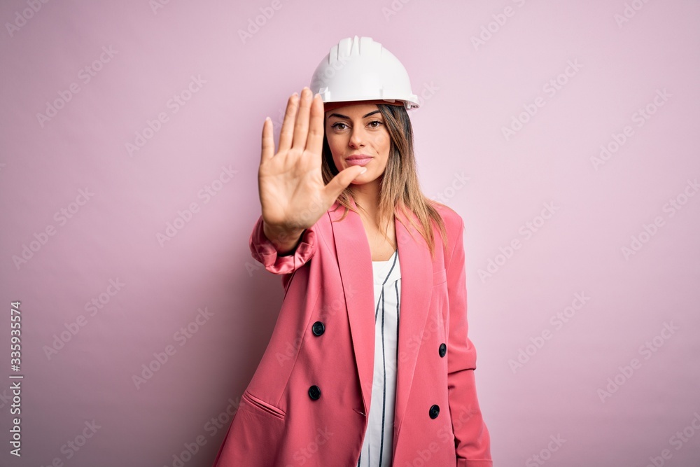 Young beautiful brunette architect woman wearing safety helmet over pink background doing stop sing with palm of the hand. Warning expression with negative and serious gesture on the face. Young beautiful brunette architect woman wearing safety helmet over pink background doing stop sing with palm of the hand. Warning expression with negative and serious gesture on the face.