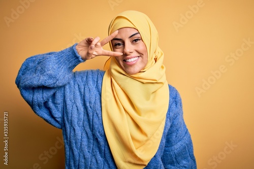 Young beautiful brunette muslim woman wearing arab hijab over isolated yellow background Doing peace symbol with fingers over face, smiling cheerful showing victory