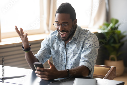 Excited African American man in glasses sit at desk triumph win online lottery on cellphone, overjoyed biracial male in spectacles feel euphoric read good unexpected news or message on smartphone