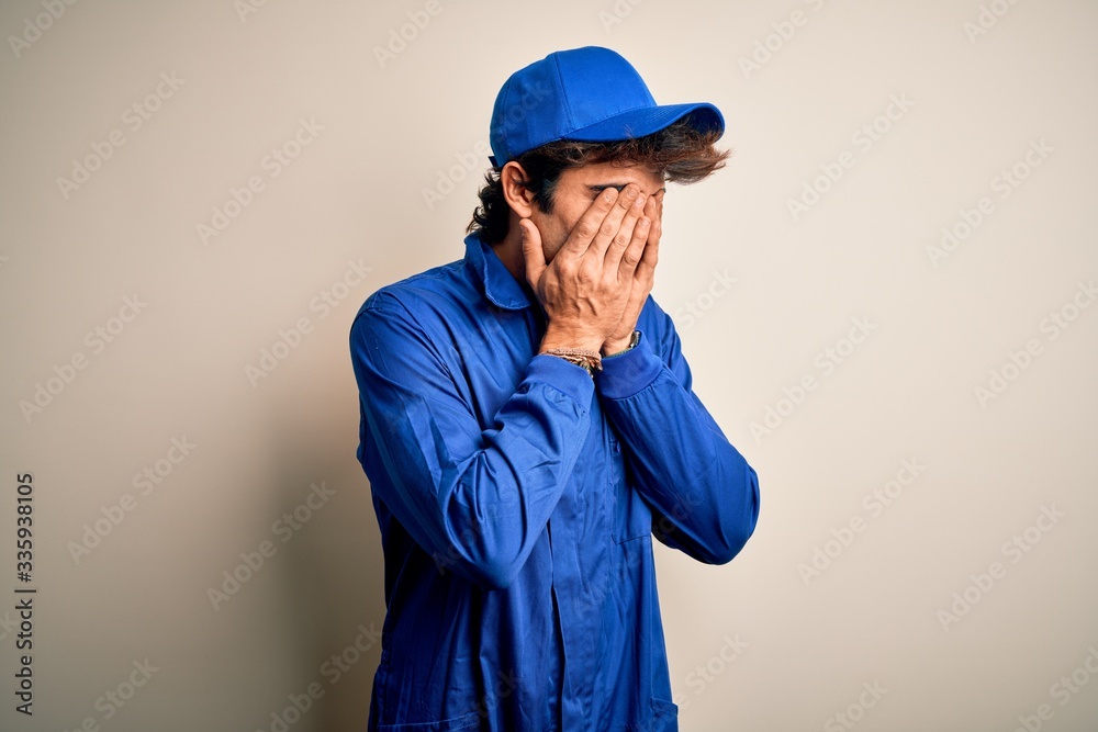 Young mechanic man wearing blue cap and uniform standing over isolated white background with sad expression covering face with hands while crying. Depression concept.