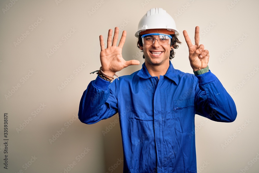 Young constructor man wearing uniform and security helmet over isolated white background showing and pointing up with fingers number seven while smiling confident and happy.