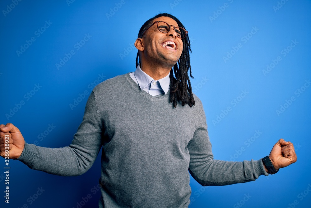 Fototapeta premium Young handsome african american man with dreadlocks wearing casual sweater and glasses very happy and excited doing winner gesture with arms raised, smiling and screaming for success. Celebration 