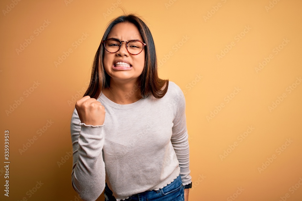 Young beautiful asian girl wearing casual sweater and glasses over yellow background angry and mad raising fist frustrated and furious while shouting with anger. Rage and aggressive concept. Young beautiful asian girl wearing casual sweater and glasses over yellow background angry and mad raising fist frustrated and furious while shouting with anger. Rage and aggressive concept.