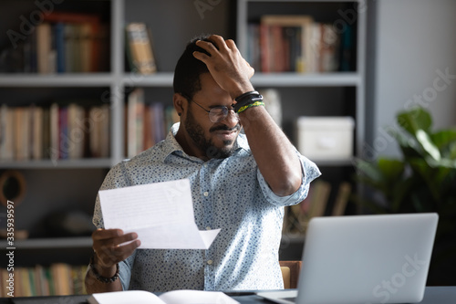 Frustrated biracial male student sit at desk feel distressed with bad news in paper letter, unhappy African American man stressed by dismissal notice or negative reply in post correspondence or notice