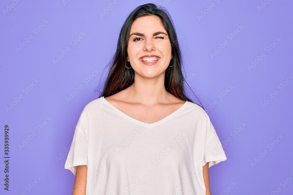 Young beautiful brunette woman wearing casual white t-shirt over purple background winking looking at the camera with sexy expression, cheerful and happy face. Young beautiful brunette woman wearing casual white t-shirt over purple background winking looking at the camera with sexy expression, cheerful and happy face.