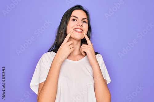 Young beautiful brunette woman wearing casual white t-shirt over purple background Smiling with open mouth, fingers pointing and forcing cheerful smile