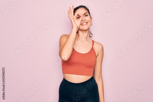 Young beautiful fitness woman wearing sport excersie clothes over pink background doing ok gesture with hand smiling, eye looking through fingers with happy face. © Krakenimages.com